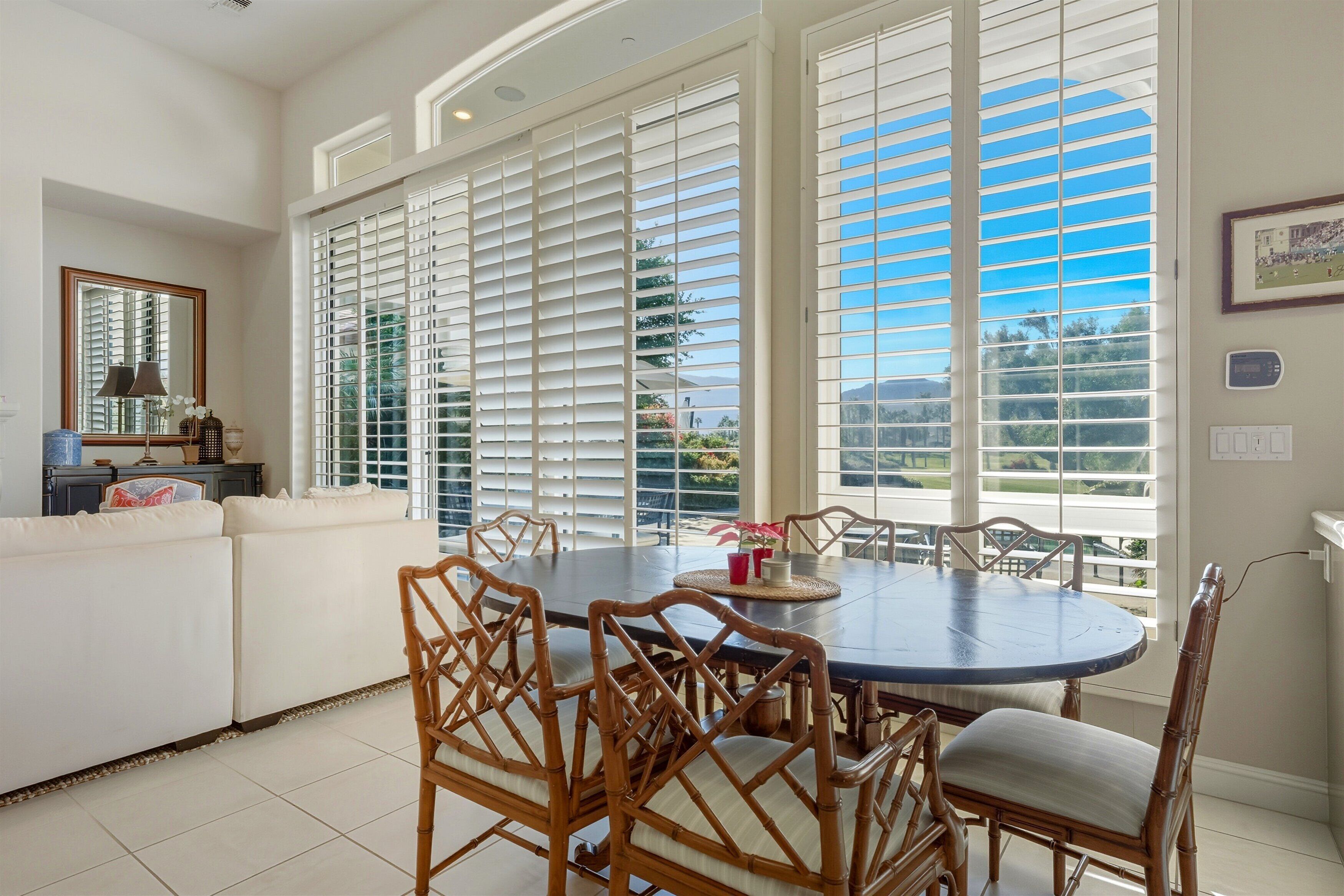 A dining area with a round table, chairs, and large windows with blinds.
