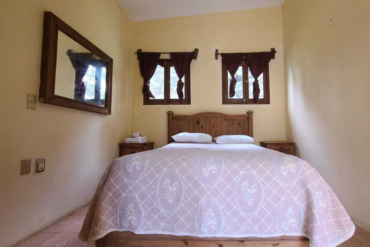 Quarto com cama, duas janelas com cortinas, mesa de cabeceira e um espelho na parede.