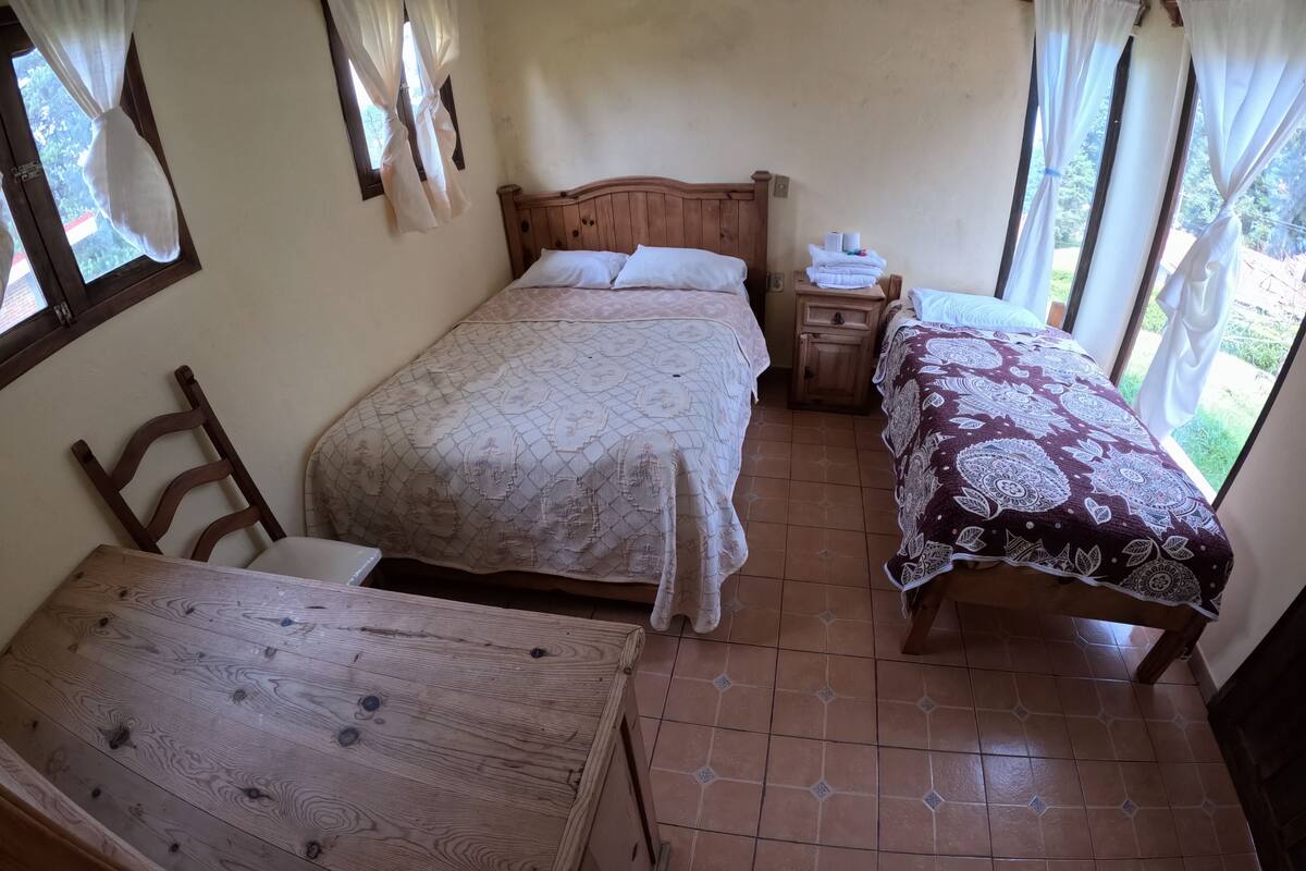 Quarto com duas camas, uma mesa de madeira e janela com cortinas.