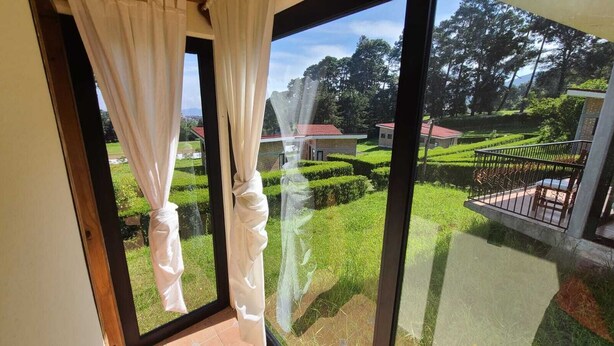 A view from a window with white curtains, overlooking a well-maintained garden with neatly trimmed hedges and a small building with a red roof. - Cabañas Entrada a la Sierra (Zacapoaxtla)