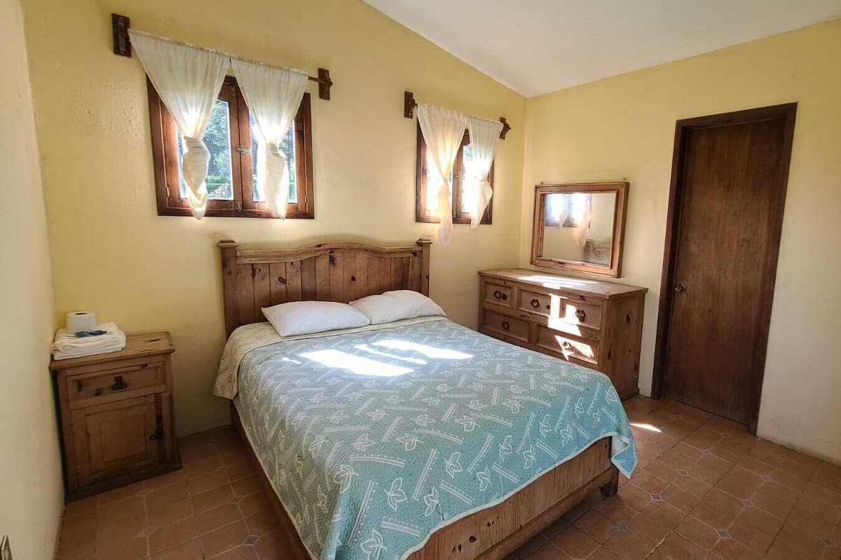 Quarto com cama de madeira, mesa de cabeceira, cômoda e porta.