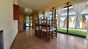 A dining area with wooden chairs and a table, large windows with curtains, and a view of a brick building and greenery outside. - Cabañas Entrada a la Sierra (Zacapoaxtla)