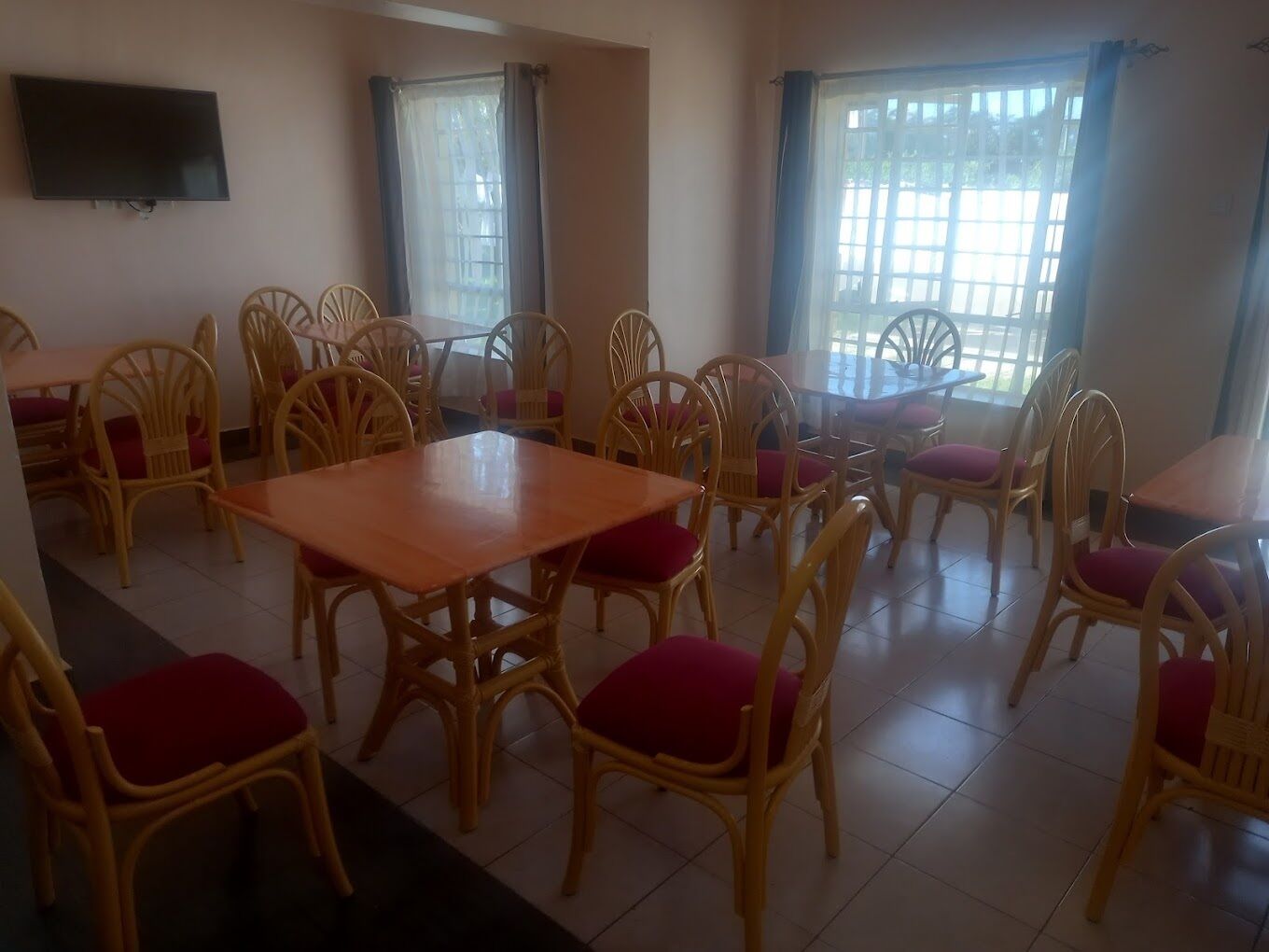A dining area with wooden tables and wicker chairs, large windows with curtains, and a flat-screen TV mounted on the wall.