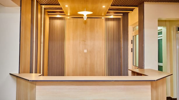 A reception area with a wooden reception desk, vertical blinds, and a ceiling light fixture. - Ack Guest House Machakos (Machakos)