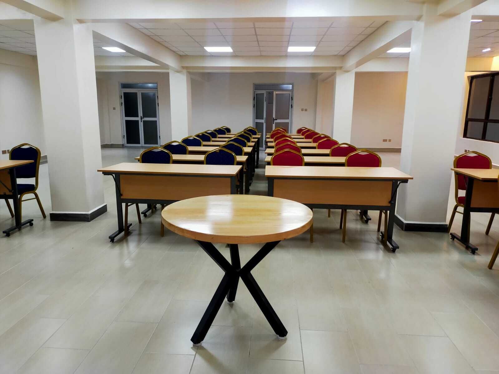 Uma sala de aula com fileiras de mesas e cadeiras, uma mesa redonda no centro e uma porta ao fundo.