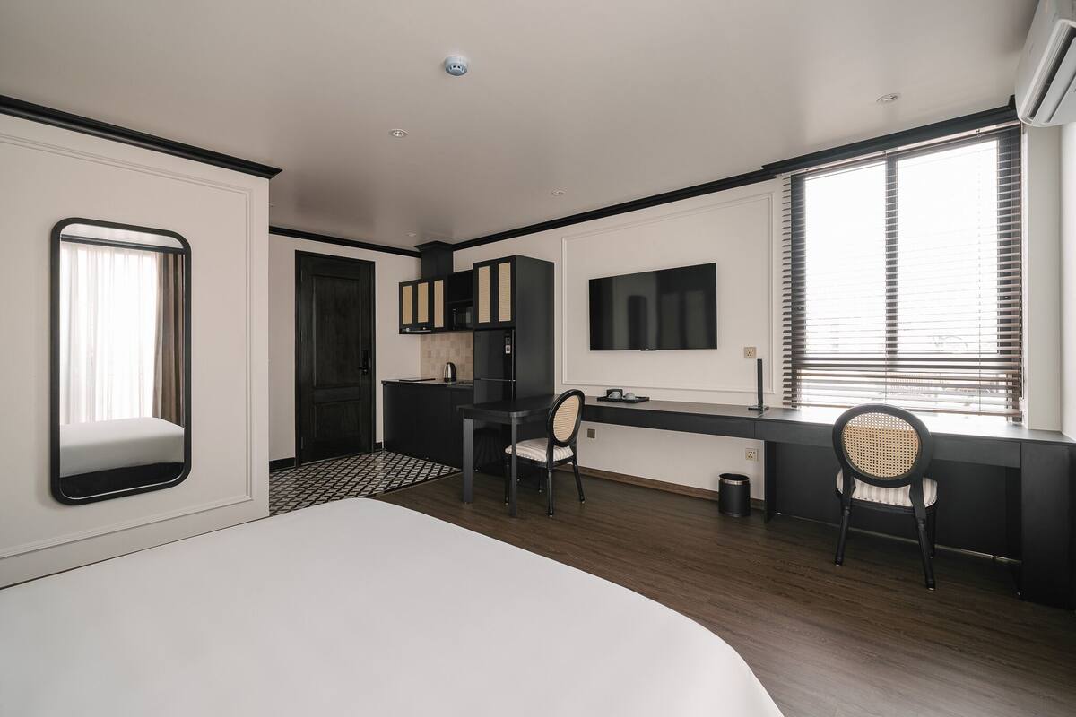 A modern hotel room with a large bed, a desk with a chair, a TV mounted on the wall, and a window with blinds.
