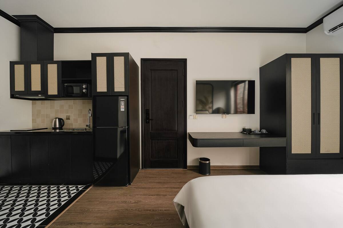 A modern hotel room with a kitchenette, a bed, a mirror, and a dark wooden door.