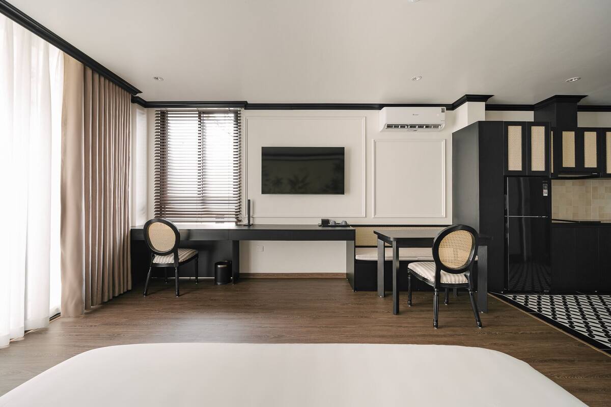 A modern hotel room with a wooden floor, a bed, a desk with two chairs, a large window with blinds, and a kitchen area with cabinets.