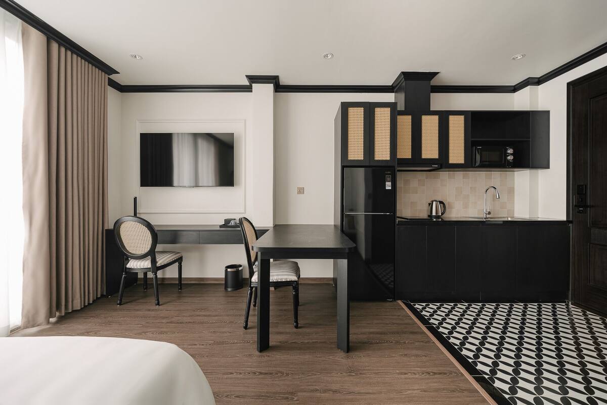 A modern hotel room with a dining area, a flat-screen TV mounted on the wall, a black kitchen with a microwave, and a patterned rug.