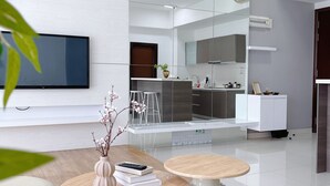 Un salon moderne doté d’un téléviseur à écran plat, de deux tables rondes, d’un canapé et d’un coin cuisine avec un grand miroir et des meubles blancs.