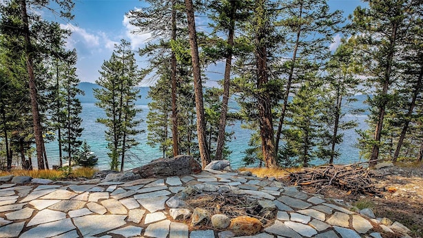 A stone-paved area with a fire pit surrounded by tall pine trees and a lake in the background. - Lakefront Home. Built 2016. For Remote work, boating, relaxing, hiking. (Marion)