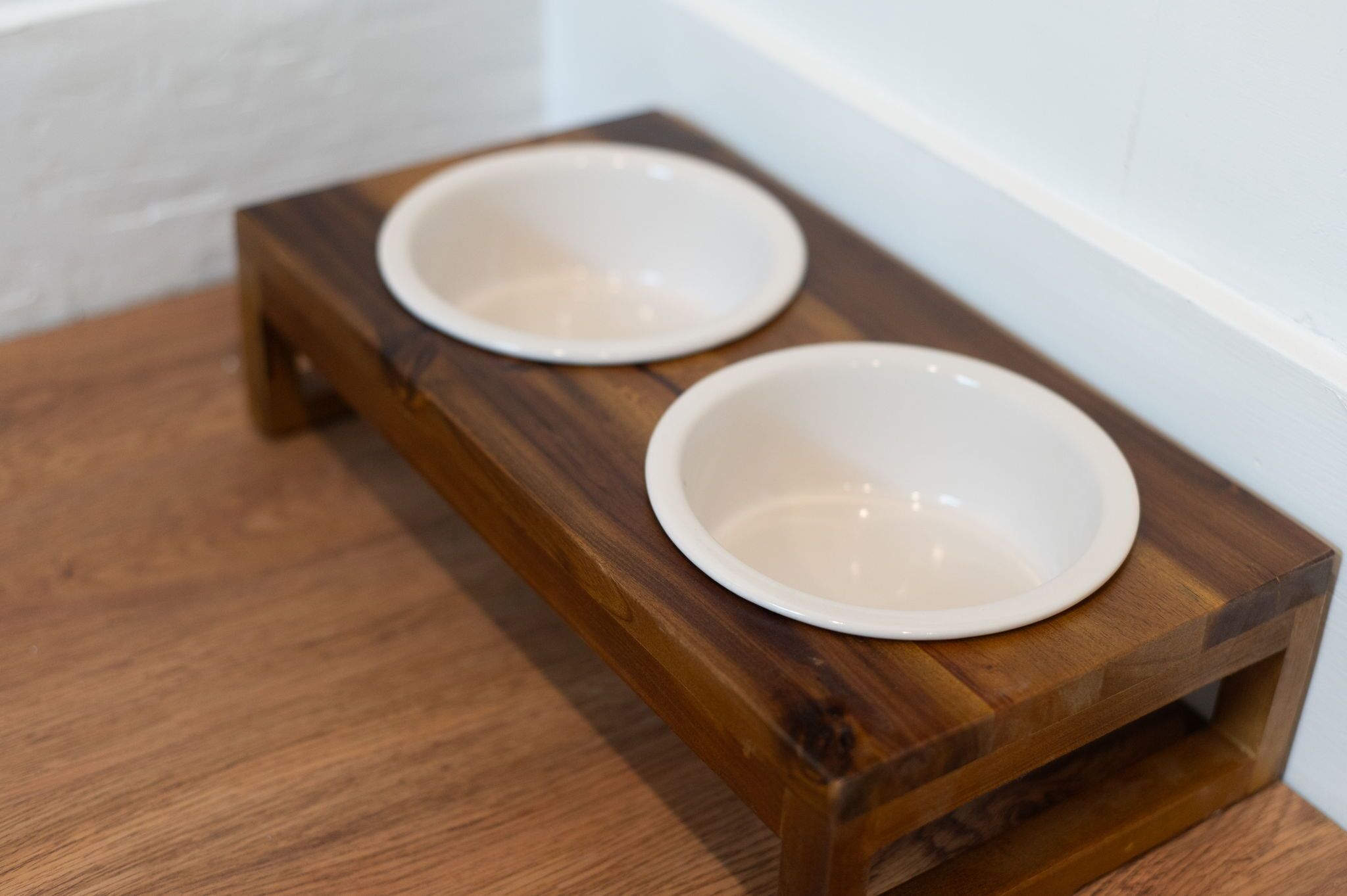 A wooden pet feeding station with two white bowls on a wooden floor.