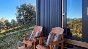 Two wooden chairs with fur throws on a wooden deck outside a building with a dark exterior and a large window.