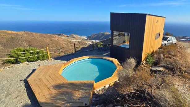 Una piscina moderna al aire libre con cubierta de madera, una pequeña estructura de madera con ventanal y vista al océano.