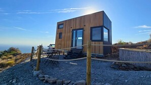 Una cabaña moderna de madera con un amplio ventanal, un comedor con mobiliario exterior y vista al océano.