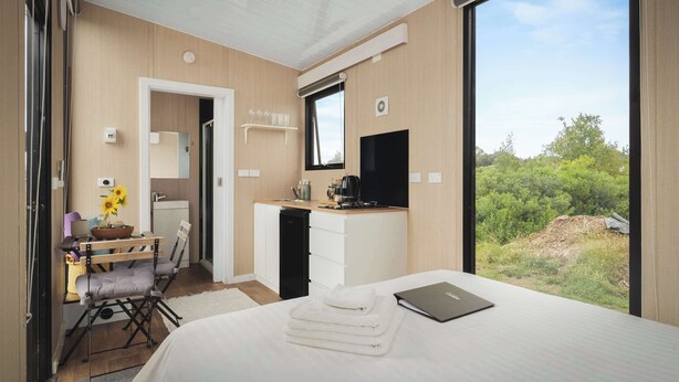 A modern hotel room with a bed, a desk with a computer, a TV, and a large window with a view of greenery.