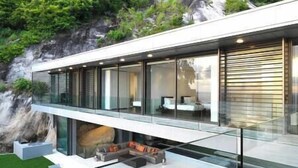 A modern house with a glass balcony, a stone wall, and a small outdoor seating area.