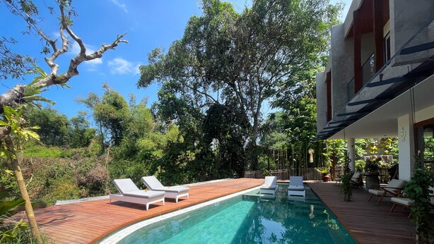 Une piscine dotée d’une terrasse en bois et de chaises longues, entourée d’une végétation luxuriante et d’arbres.