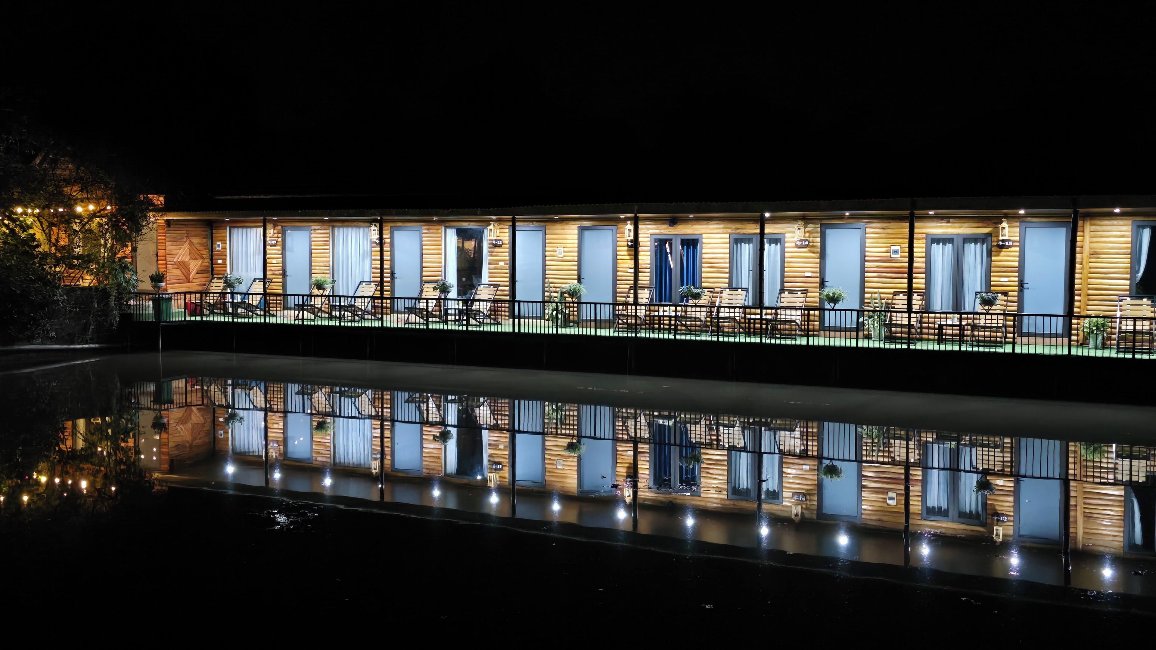 A row of log cabin-style rooms with large windows, a pool in the foreground, and a reflection of the building.
