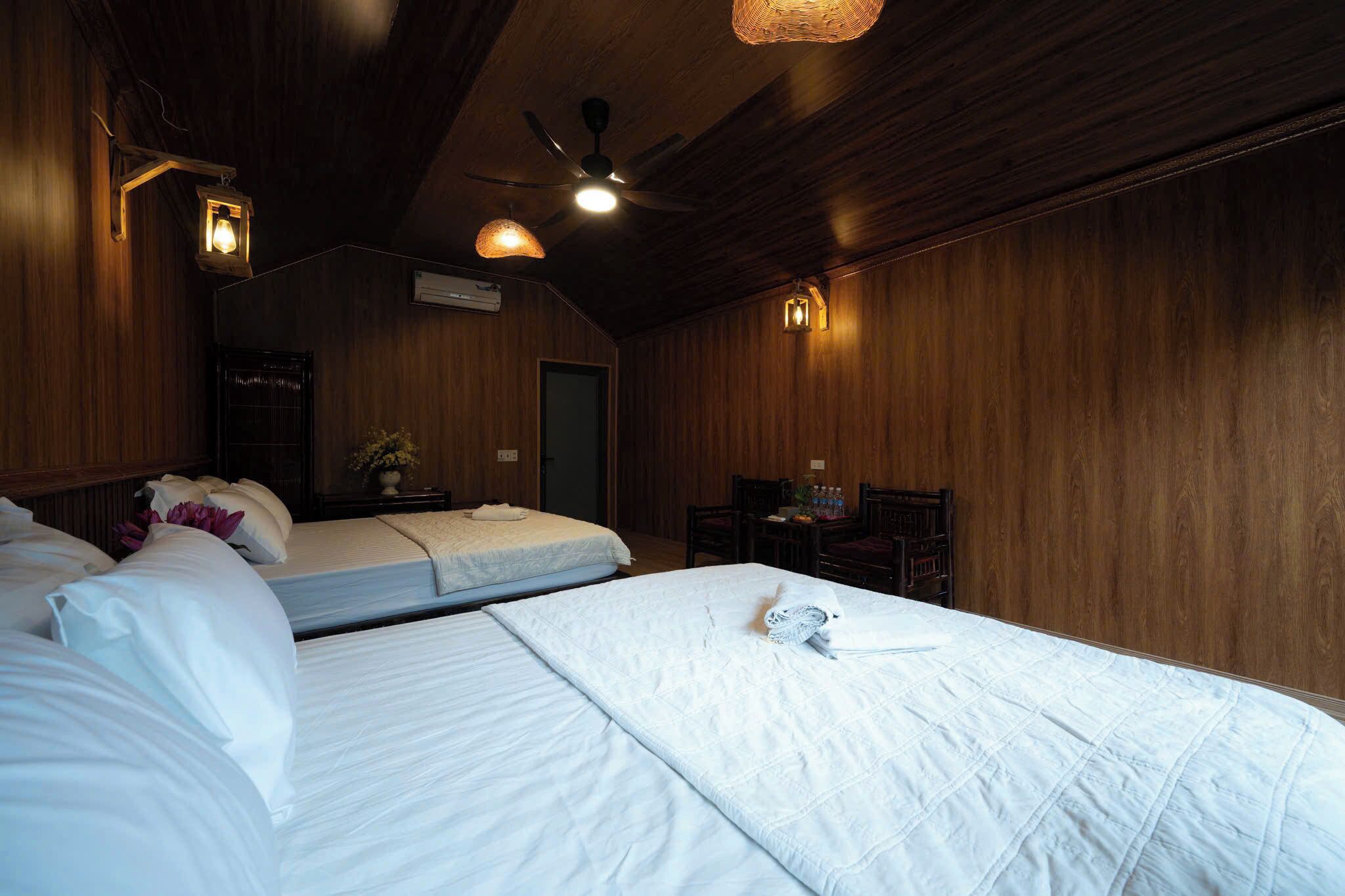 A hotel room with wooden walls, two beds, a ceiling fan, and a small table with a chair.
