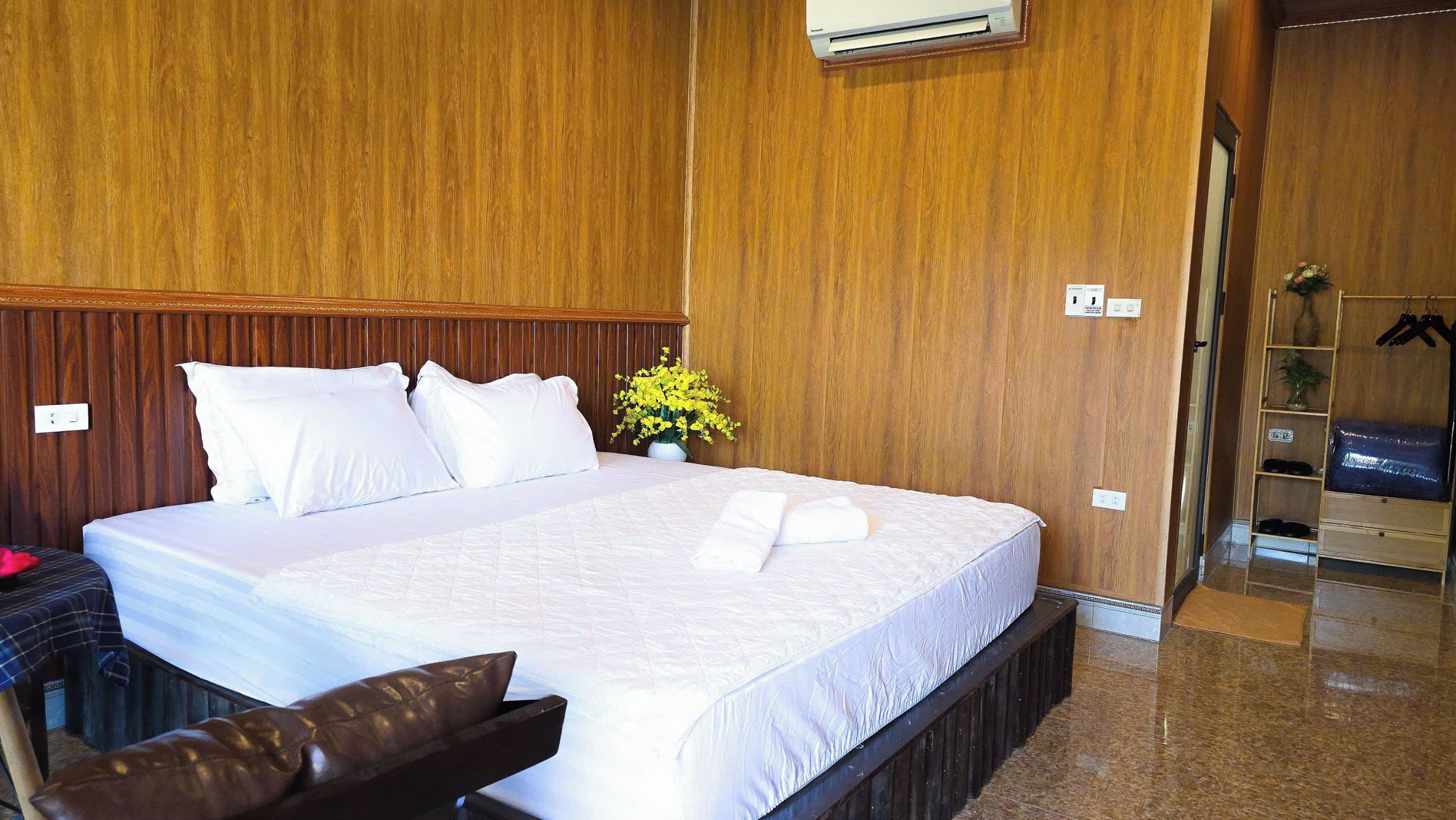 A hotel room with a large bed, wooden paneling on the walls, a small table with a vase of flowers, and a wardrobe with hanging clothes.