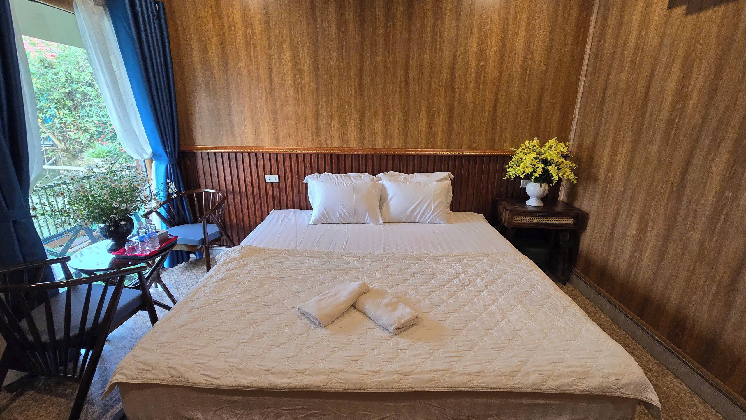 A neatly made bed with pillows and a folded towel, a wooden headboard, a small table with a chair, and a window with curtains.