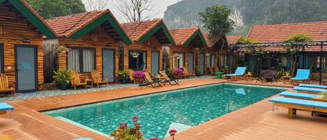 A pool area with wooden deck, lounge chairs, and cabins with red roofs.