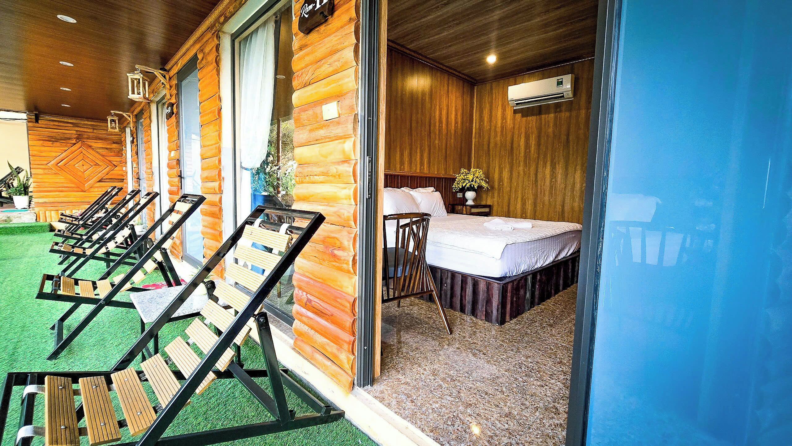 A room with a bed, a small table, and a chair. There are also several lounge chairs outside the room.