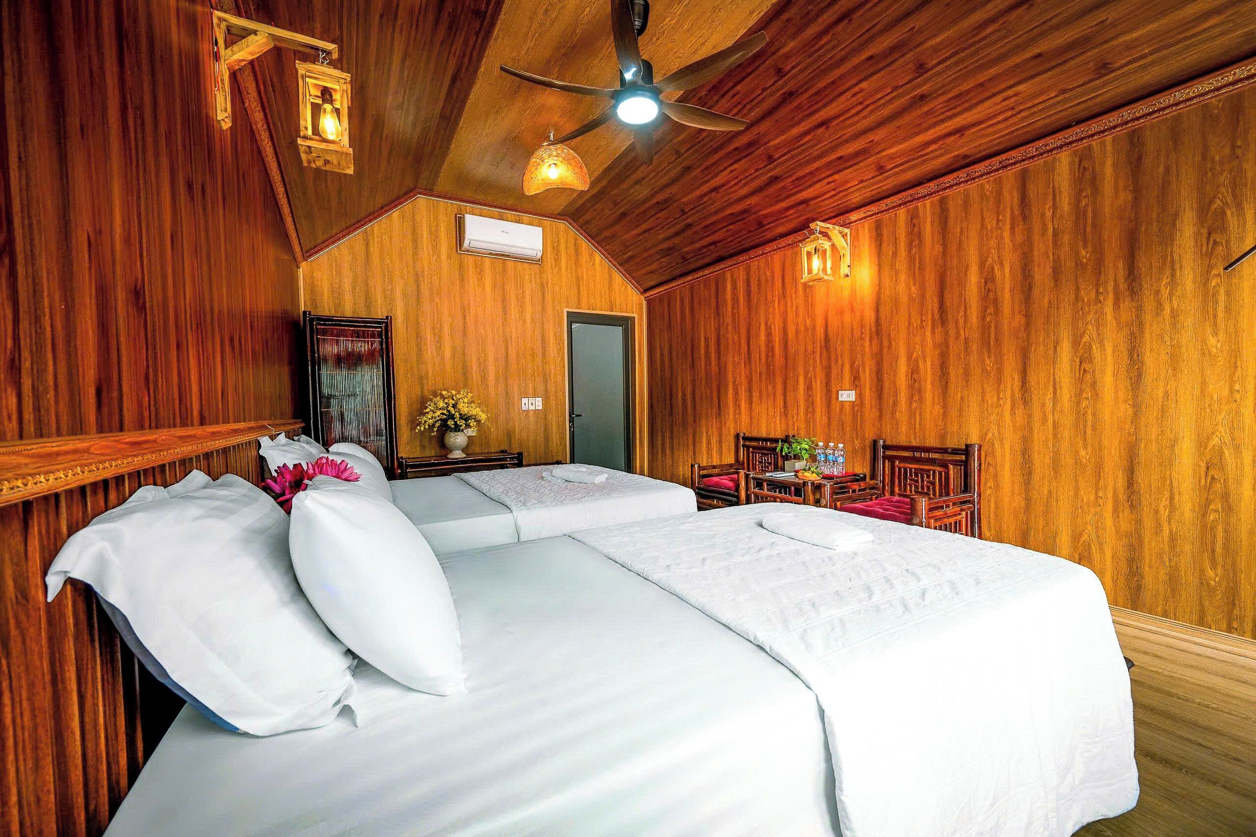 Una habitación con paredes de madera, un ventilador de techo, una cama con sábanas blancas, una mesa pequeña con sillas y una puerta.