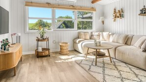 A cozy living room with a wooden ceiling, white walls, a large window with a view, a sofa, a coffee table, and a TV on a wooden stand.