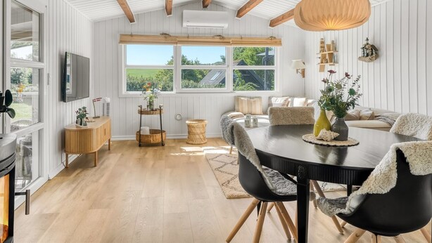 A modern dining area with a wooden ceiling, a round table, and chairs. There are windows providing natural light and a view of greenery outside.