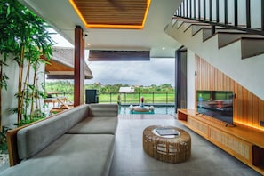 A modern living room with a sofa, a TV, a pool, and a view of the outdoors. - Kairos Villa Ubud by Omkara Experience (Ubud)