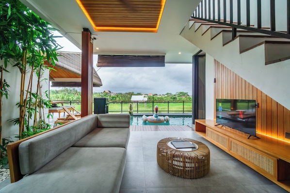 A modern living room with a sofa, a TV, a pool, and a view of the outdoors.