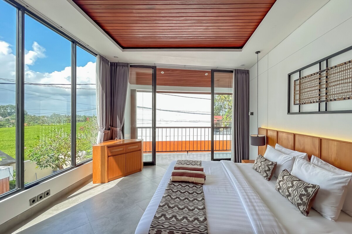 A spacious bedroom with a large bed, a wooden headboard, a nightstand, a desk, and a view of a green landscape through large windows.