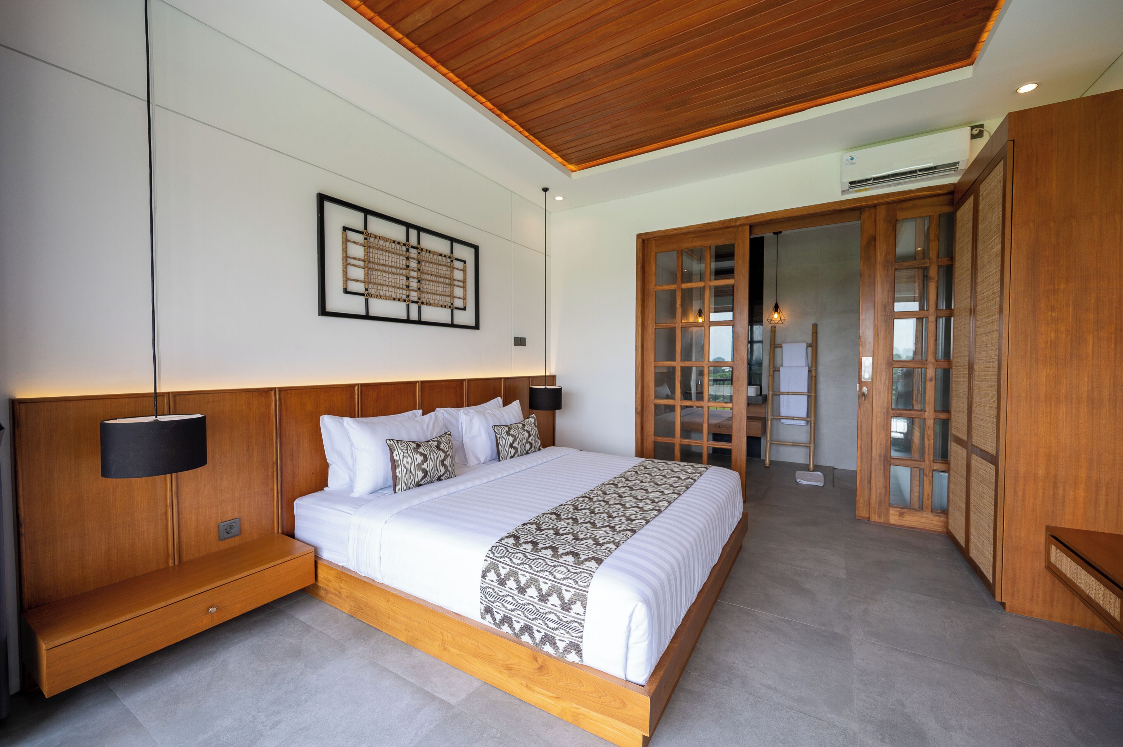 Un dormitorio moderno con cabecera de madera, cama grande, mesita de noche, una obra de arte en la pared y puerta corrediza de cristal que da a un balcón.