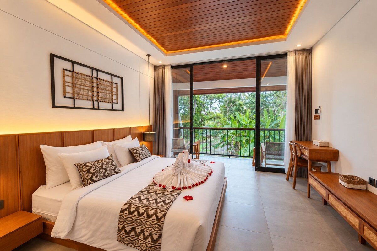 A hotel room with a wooden ceiling, a large bed with white bedding, a desk, and a balcony with a view of greenery.