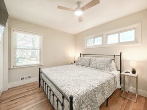 Une chambre à coucher avec un lit, un ventilateur de plafond, deux fenêtres, une table de chevet et une petite plante.