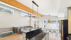 A modern kitchen with a central island, wooden ceiling beams, and a view of a body of water through large windows. - Oceanfront w/ Incredible Views (Juneau)