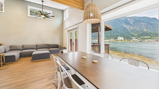 A modern living room with a wooden ceiling, a large dining table, and a view of a lake and mountains. - Oceanfront w/ Incredible Views (Juneau)
