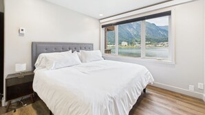 A bedroom with a large bed, a nightstand, a lamp, and a window with a mountain view. - Oceanfront w/ Incredible Views (Juneau)