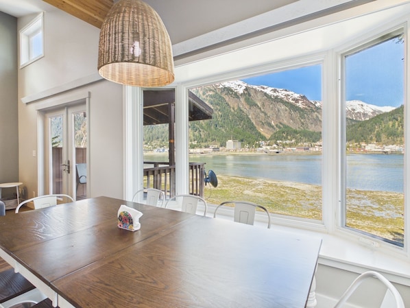 Ein Essbereich mit Holztisch, Stühlen, einer Hängeleuchte und großen Fenstern mit Blick auf einen See und Berge.