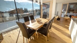 Et moderne spiseområde med et træbord og sorte stole, store vinduer med udsigt til bjerge, og et køkken med træskabe.