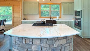 Una cocina con una isla grande, base de piedra, armarios verde claro y una chimenea de piedra.