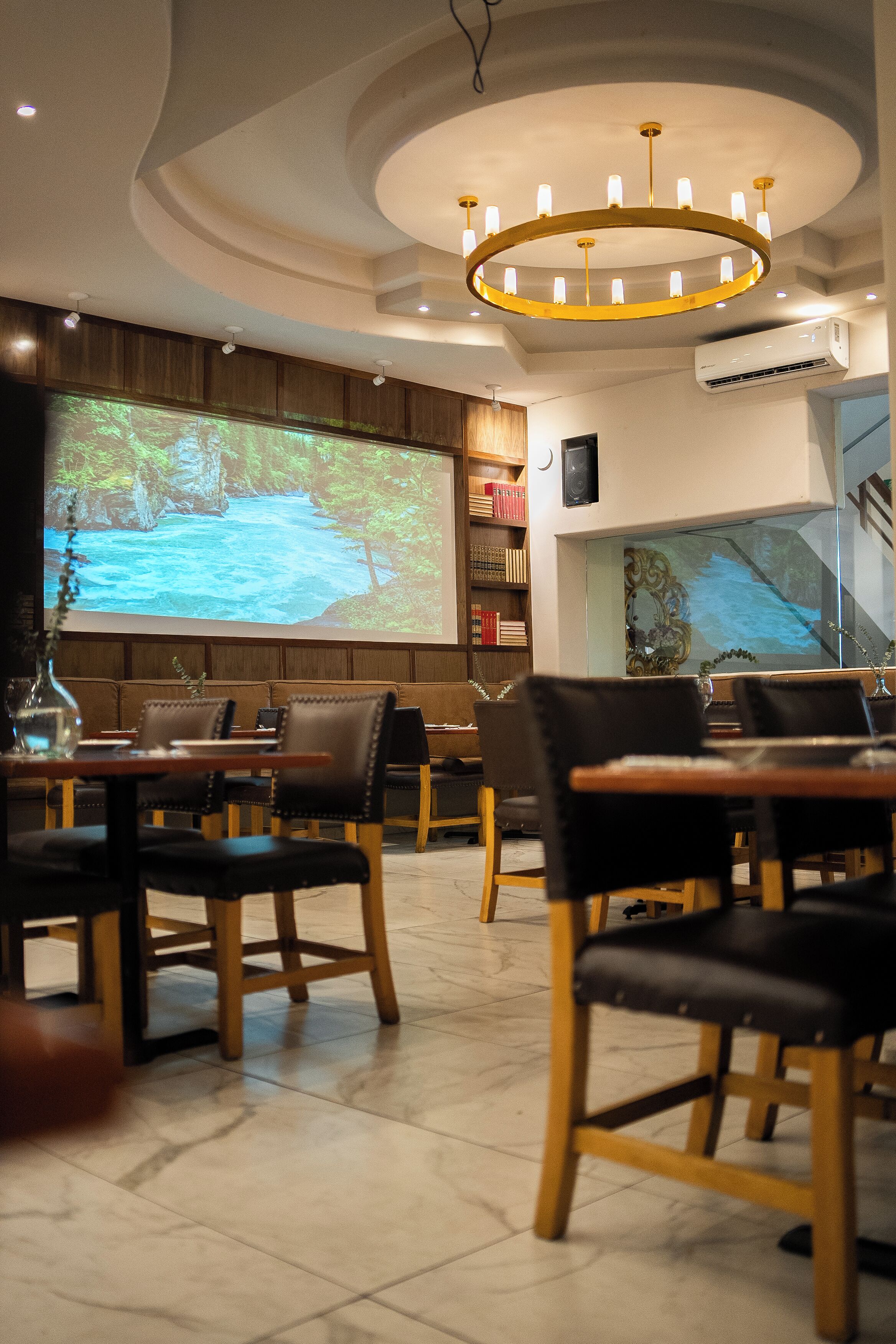 Een restaurantinterieur met een projectiescherm waarop een natuurscène te zien is, houten meubels en een moderne kroonluchter.