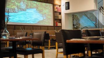 Een restaurantinterieur met een projectiescherm waarop een natuurscène te zien is, houten meubels en een moderne kroonluchter.