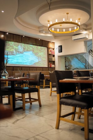 Een restaurantinterieur met een projectiescherm waarop een natuurscène te zien is, houten meubels en een moderne kroonluchter.