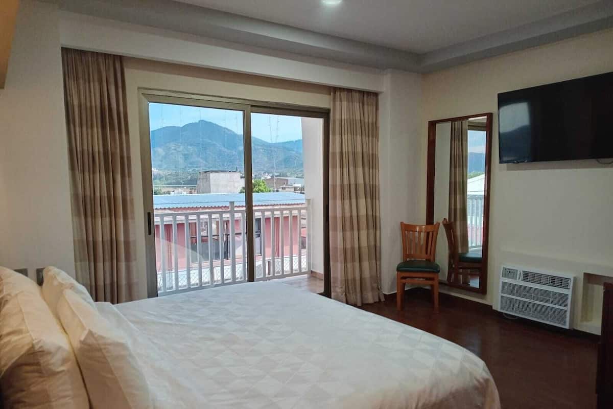 A hotel room with a large bed, a chair, a TV mounted on the wall, and a balcony with curtains.