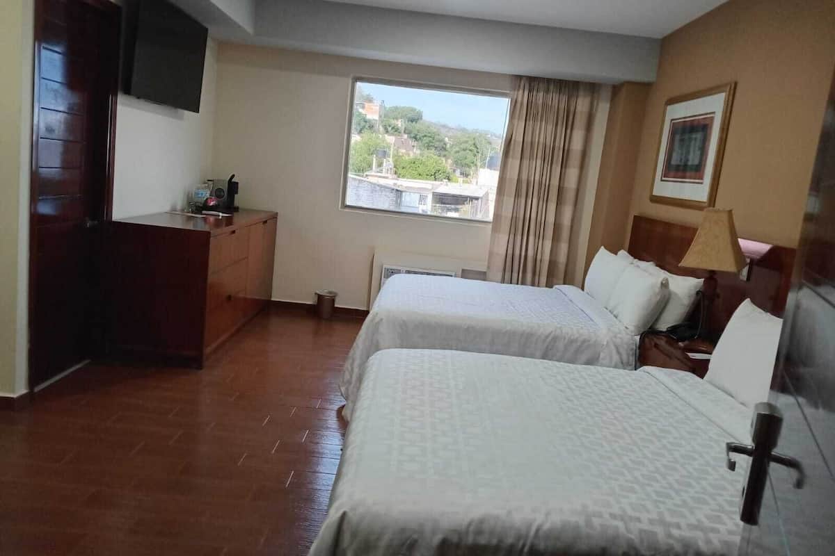 A hotel room with two beds, a desk, a TV, a window with a view, and a lamp.