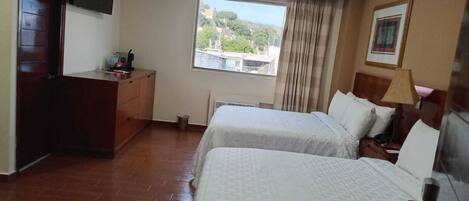 A hotel room with two beds, a desk, a TV, a window with a view, and a lamp.