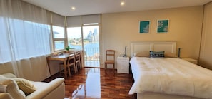 A hotel room with a bed, a desk, a chair, a nightstand, and a view of a cityscape through large windows.